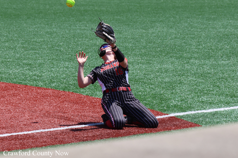 Softball fielder in pinstriped uniform dives on red dirt, glove raised to catch an airborne ball on a green field nearby.