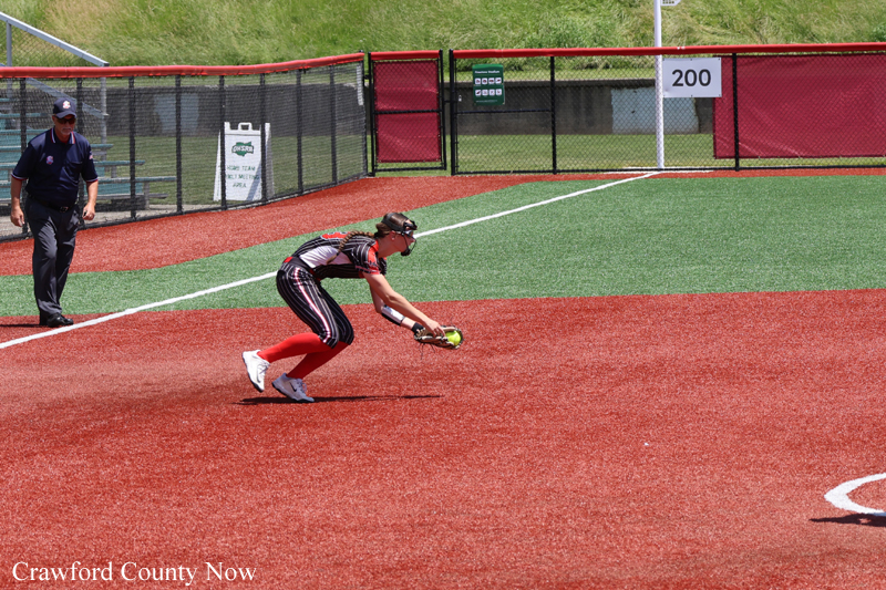 Softball catcher dives to field a ground ball on a red infield while an umpire watches nearby.