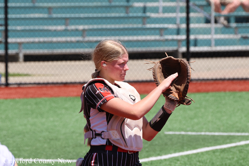 Young female softball catcher in white chest protector raises a glove, ready to catch on a sunny field with bleachers behind her.