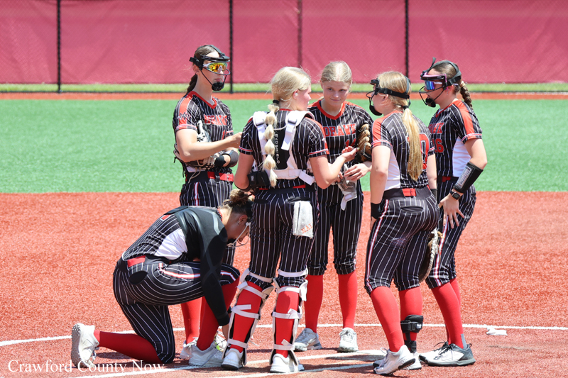 Girls softball team huddles on the infield in black pinstripe uniforms with red socks, one kneeling catcher adjusting gear as teammates talk nearby on a red dirt field.