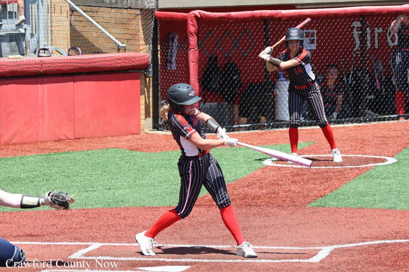 Batter in a black pinstripe uniform swings a bat on a softball field, wearing a helmet and red socks, with a fencer behind infield dirt and green turf nearby.