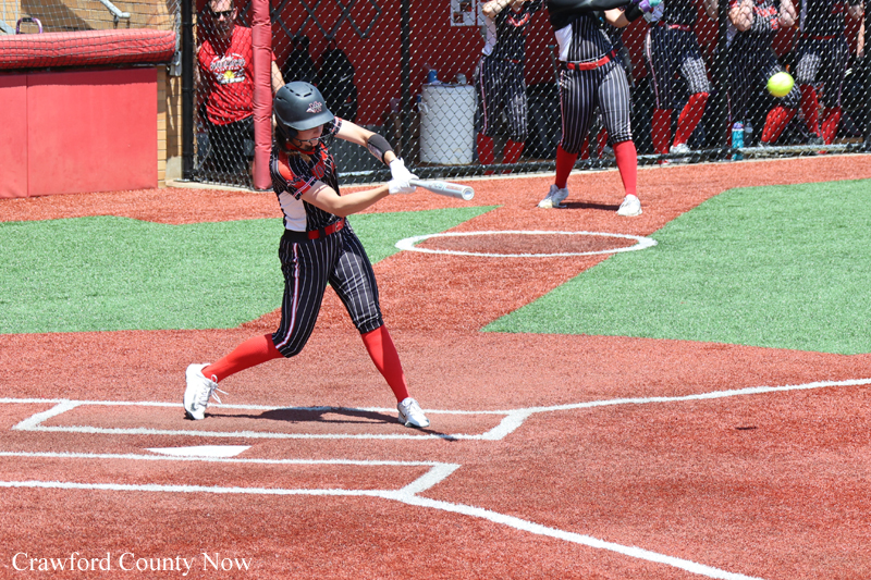 Young female softball batter in a striped uniform swings at a pitch near home plate with teammates and fans in the dugout behind her on a dirt field.