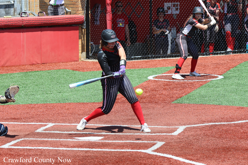 Female softball batter in black jersey and pinstriped pants swings at a pitch at home plate on a red-dirt field with red socks and helmet.