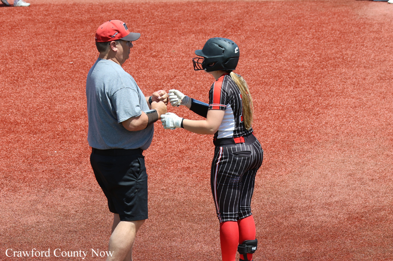 Adult coach in a red cap fist-bumps a female softball catcher in a black pinstripe uniform and helmet on a red dirt field.