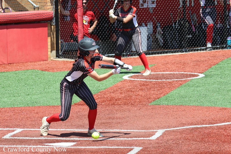 Young female softball batter in a black pinstripe uniform swings at a pitch at home plate during a game in progress.