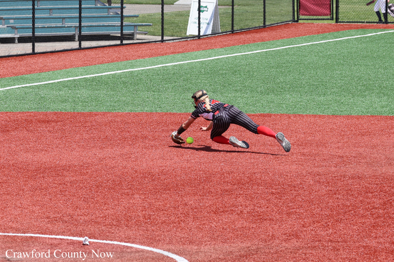 Diving softball infielder reaches for a ground ball on a red dirt infield with green outfield behind her.