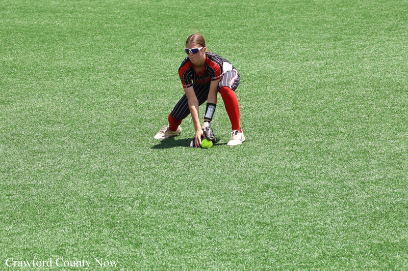 A female softball catcher crouches on a green field, wearing protective gear and sunglasses, placing a glove near a ball on the ground.