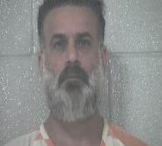 Mugshot of a middle-aged man with a gray beard in an orange-and-white striped prison jumpsuit, standing against a gray wall; timestamp shown.