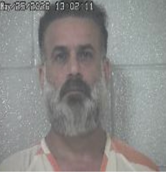 Mugshot of a middle-aged man with a gray beard in an orange-and-white striped prison jumpsuit, standing against a gray wall; timestamp shown.