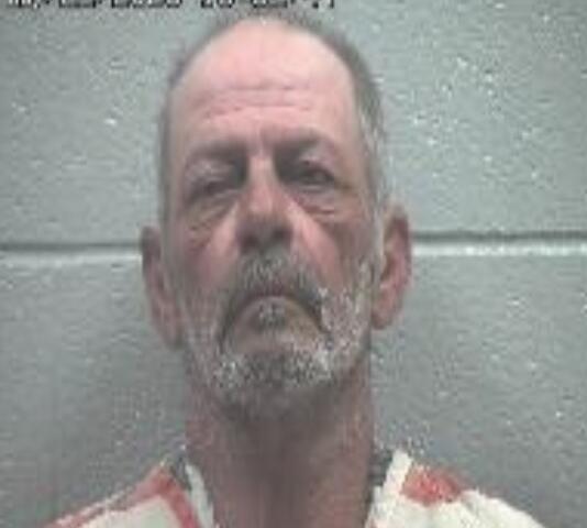 Mugshot of a middle-aged man with a grey beard in a striped jail uniform, against a grey wall; timestamp reads 10:53:07.