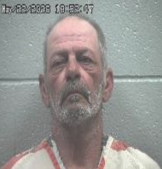 Mugshot of a middle-aged man with a grey beard in a striped jail uniform, against a grey wall; timestamp reads 10:53:07.