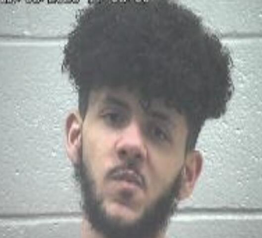 Mugshot-style photo of a young man with dark curly hair and beard against a gray wall, facing camera.