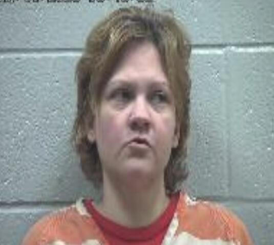 Mugshot of a woman with short brown hair wearing an orange jail uniform against a gray cinder block wall.