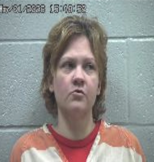 Mugshot of a woman with short brown hair wearing an orange jail uniform against a gray cinder block wall.
