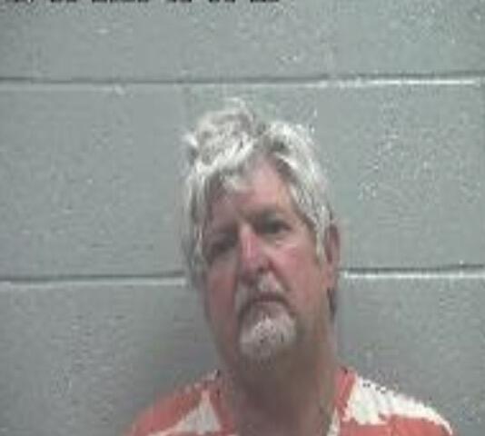 Mugshot of a middle-aged man with gray hair and a goatee wearing an orange prison jumpsuit, standing against a gray cinder block wall; a date stamp is visible in the top-left corner.