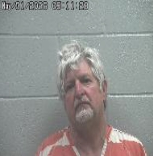 Mugshot of a middle-aged man with gray hair and a goatee wearing an orange prison jumpsuit, standing against a gray cinder block wall; a date stamp is visible in the top-left corner.