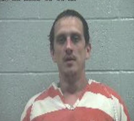 Mugshot of a man in an orange-and-white striped jail jumpsuit against a gray cinder block wall, neutral expression.