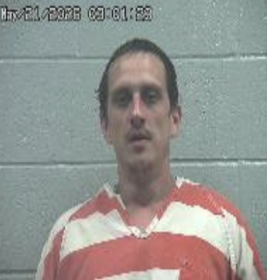 Mugshot of a man in an orange-and-white striped jail jumpsuit against a gray cinder block wall, neutral expression.