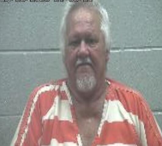 Mugshot of an older man in an orange-and-white striped prison jumpsuit standing against a gray cinder block wall with a date stamp.