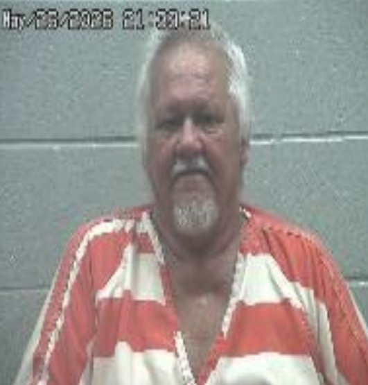Mugshot of an older man in an orange-and-white striped prison jumpsuit standing against a gray cinder block wall with a date stamp.