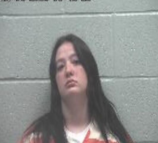 Mugshot of a young woman with long dark hair in an orange jumpsuit against a gray cinder block wall.