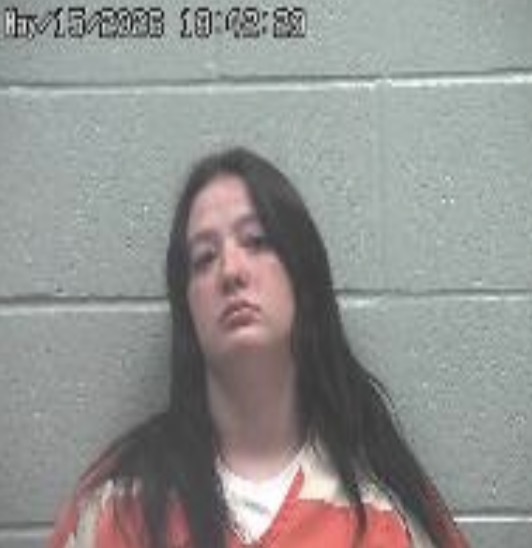 Mugshot of a young woman with long dark hair in an orange jumpsuit against a gray cinder block wall.