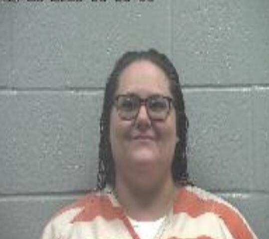 Mugshot of a woman with glasses in an orange-and-white striped jail jumpsuit, against a gray cinder block wall; timestamp in upper left.