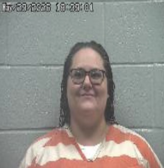 Mugshot of a woman with glasses in an orange-and-white striped jail jumpsuit, against a gray cinder block wall; timestamp in upper left.