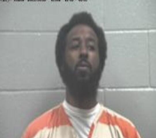 Mugshot of a man with a beard wearing an orange jail jumpsuit, standing against a gray cinder-block wall; timestamp 12:10:36 in the upper-left corner.
