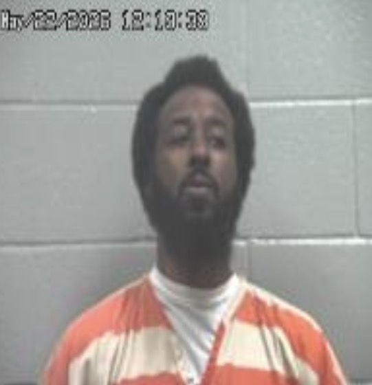 Mugshot of a man with a beard wearing an orange jail jumpsuit, standing against a gray cinder-block wall; timestamp 12:10:36 in the upper-left corner.