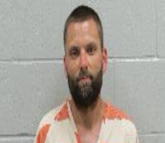 Mugshot: bearded man in an orange-and-white striped prison jumpsuit against a gray wall.