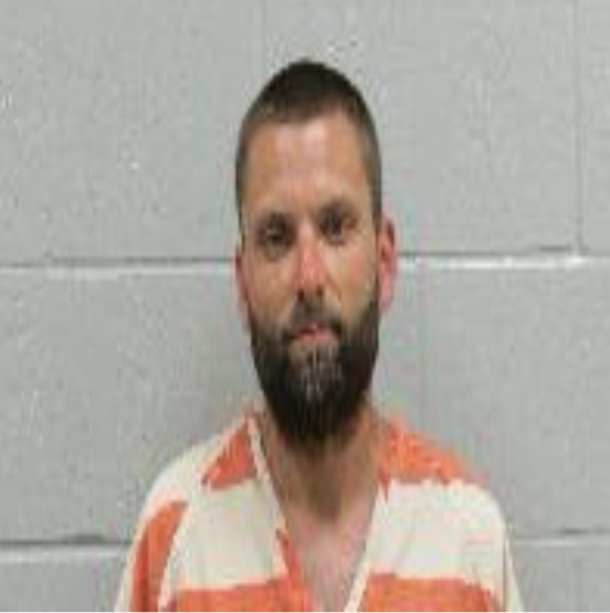 Mugshot: bearded man in an orange-and-white striped prison jumpsuit against a gray wall.