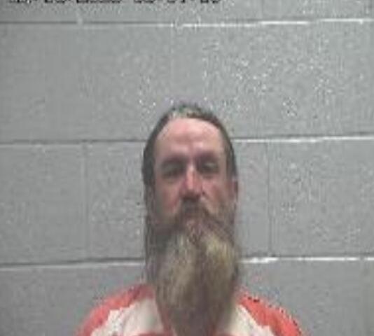 Mugshot of a bearded man in an orange-and-white striped jail uniform against a gray cinder block wall; timestamp visible in the upper-left.