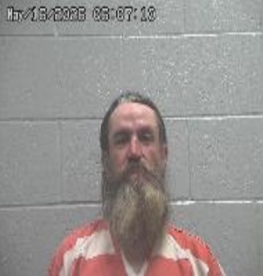 Mugshot of a bearded man in an orange-and-white striped jail uniform against a gray cinder block wall; timestamp visible in the upper-left.