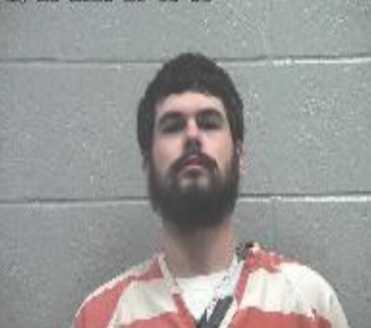 Mugshot of a bearded man in an orange jail uniform against a gray cinder-block wall, with a timestamp overlay in the upper left.