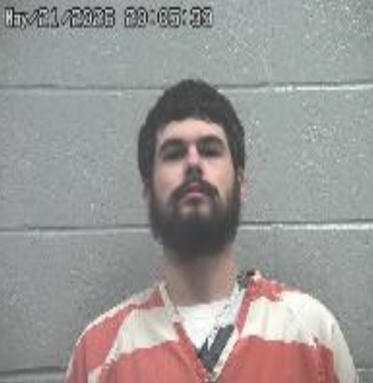 Mugshot of a bearded man in an orange jail uniform against a gray cinder-block wall, with a timestamp overlay in the upper left.