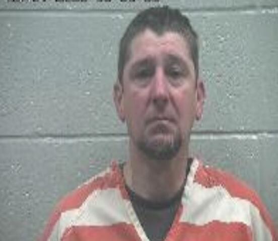 Mugshot of a middle-aged man in an orange-and-white prison jumpsuit standing in front of a gray cinder block wall, with a timestamp at the top edge.