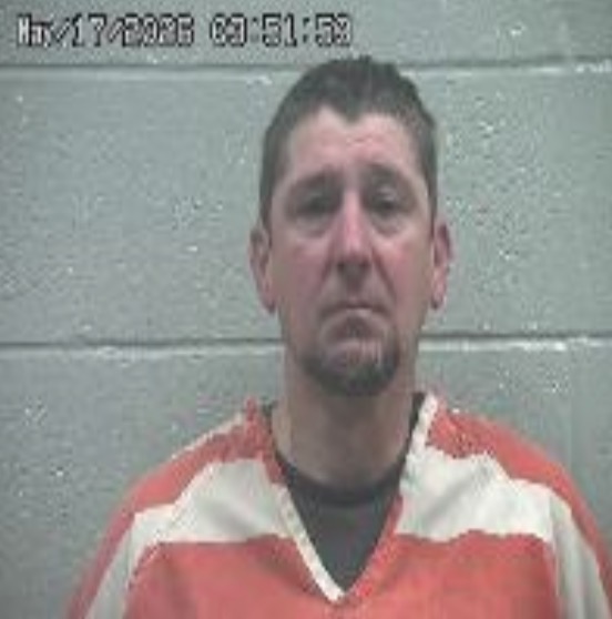 Mugshot of a middle-aged man in an orange-and-white prison jumpsuit standing in front of a gray cinder block wall, with a timestamp at the top edge.