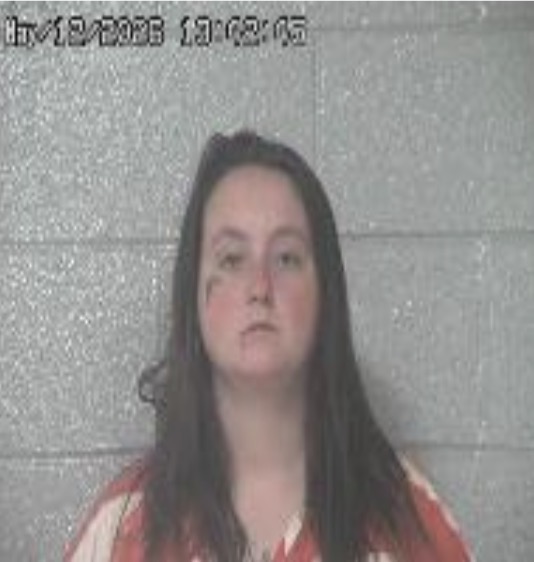Mugshot of a woman with long dark hair, wearing a red-and-white striped shirt, against a gray cinder block wall.