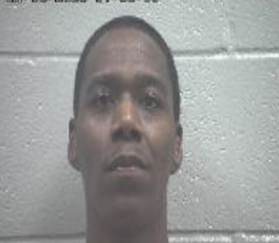 Mugshot of a man looking straight ahead against a gray cinder block wall; a date/time stamp is visible in the upper left corner.