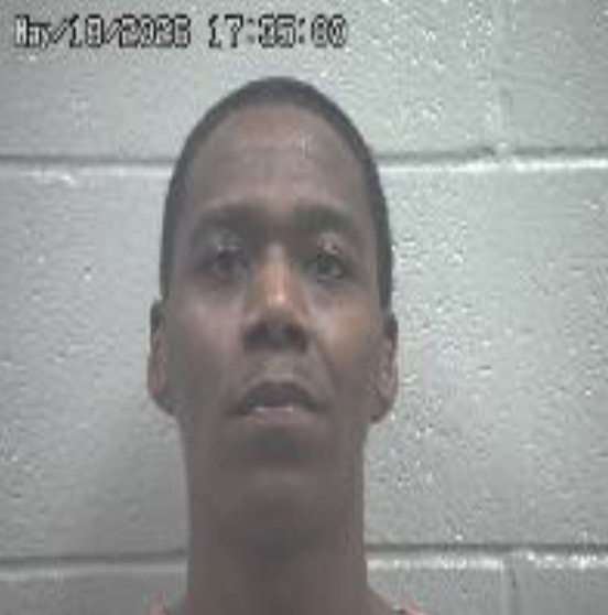 Mugshot of a man looking straight ahead against a gray cinder block wall; a date/time stamp is visible in the upper left corner.