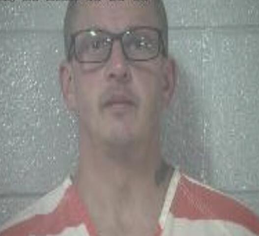 Mugshot of a bald man with glasses in a red-and-white striped prison uniform, standing against a gray wall; timestamp visible at top.