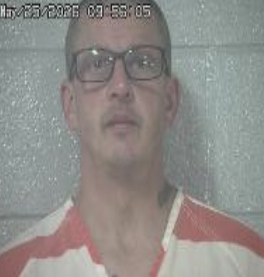 Mugshot of a bald man with glasses in a red-and-white striped prison uniform, standing against a gray wall; timestamp visible at top.