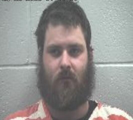 Mugshot of a bearded man in an orange-and-white striped jail uniform, looking at the camera against a gray cinder block wall.