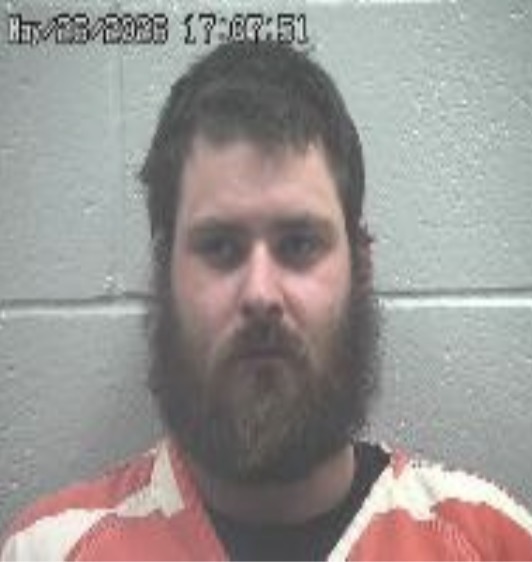Mugshot of a bearded man in an orange-and-white striped jail uniform, looking at the camera against a gray cinder block wall.