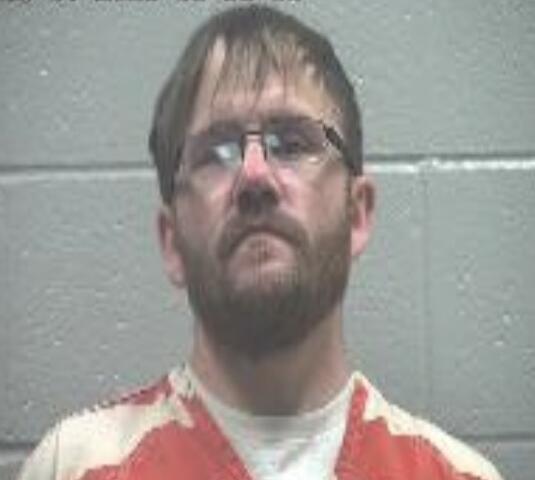 Mugshot of a bearded man wearing an orange-and-white striped jail jumpsuit, standing against a gray cinderblock wall; timestamp visible in the upper-left corner.