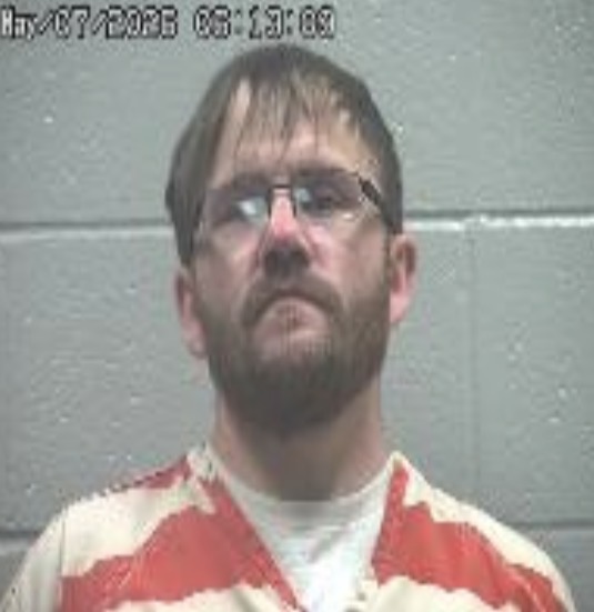 Mugshot of a bearded man wearing an orange-and-white striped jail jumpsuit, standing against a gray cinderblock wall; timestamp visible in the upper-left corner.