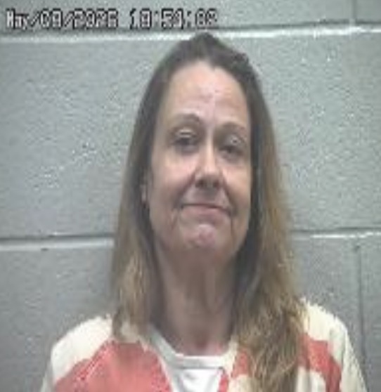 Mugshot of a middle-aged woman with shoulder-length hair in an orange-and-white striped jail uniform, against a gray cinder block wall.