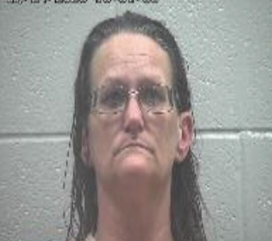 Mugshot of a middle-aged man with long dark hair and glasses, wearing an orange jumpsuit, against a grey cinder block wall with a timestamp at the top left