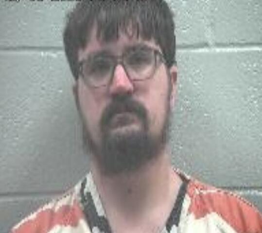 Mugshot of a man with glasses and a dark beard wearing an orange prison jumpsuit, standing against a gray cinder block wall; timestamp appears in the upper left.